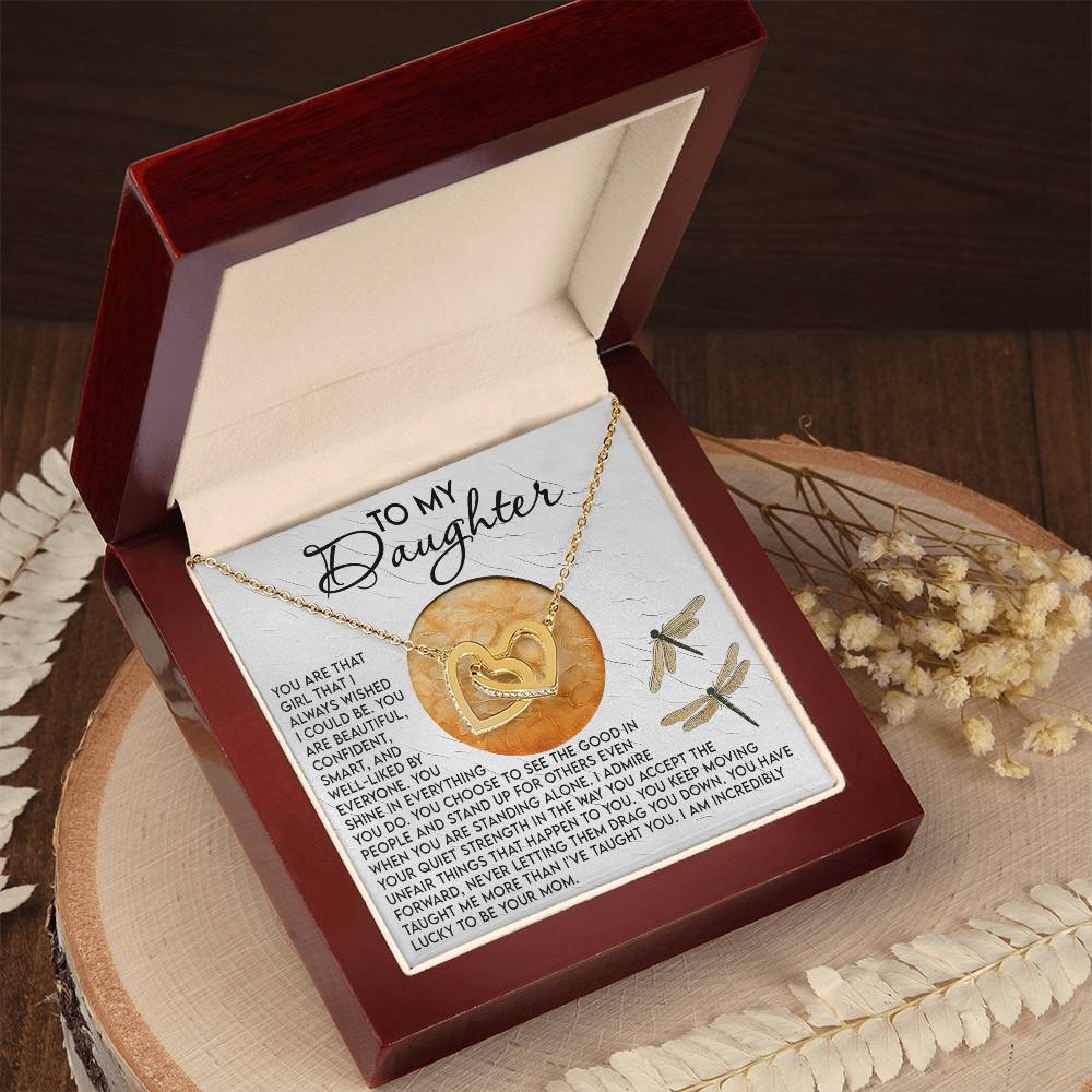 The Daughter-Quiet Strength interlocking hearts necklace, adorned with cubic zirconia crystals, is presented in a brown jewelry box featuring the message "To My Daughter." Dried flowers are scattered on the wooden surface nearby.
