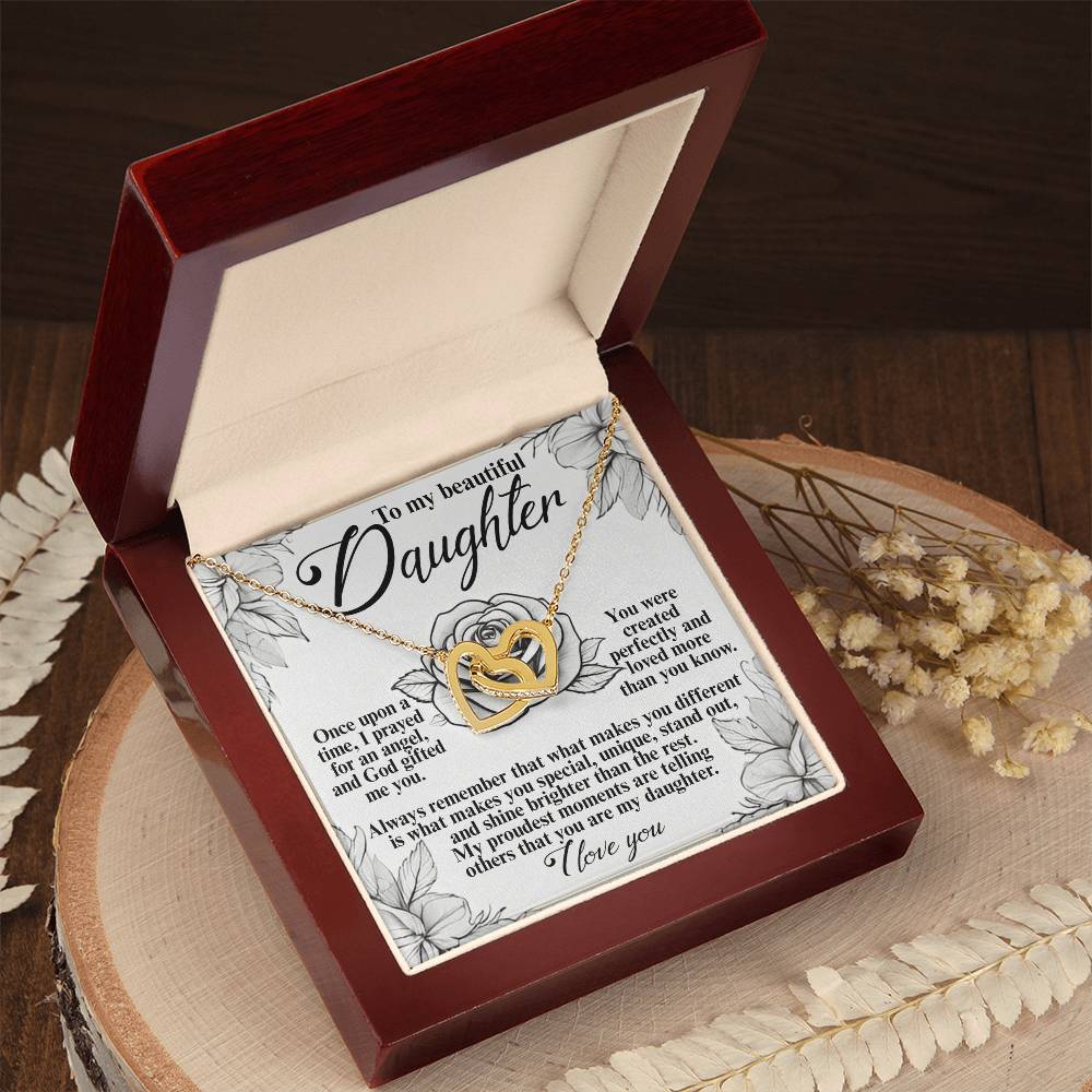 The Daughter-Prayed Angel - Interlocking Hearts Necklace, featuring cubic zirconia and a rose gold finish, rests in a red velvet box on a wooden surface. A heartfelt note for a daughter accompanies it, with dried flowers nearby for decoration.