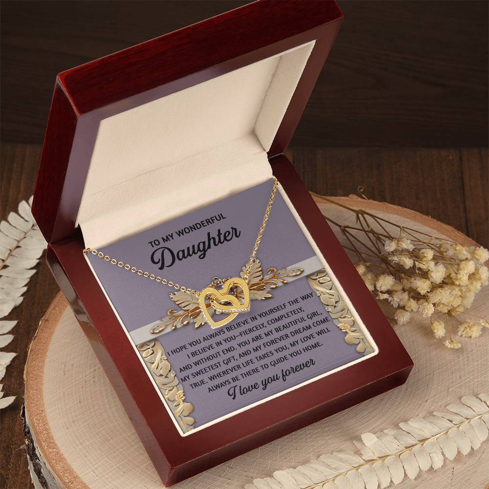 The "Daughter-Guide You Home - Interlocking Hearts Necklace," featuring a gold heart with zirconia crystals, is showcased in an open red and cream jewelry box on a wooden surface with dried flowers. A message for a daughter is partially visible inside the box.