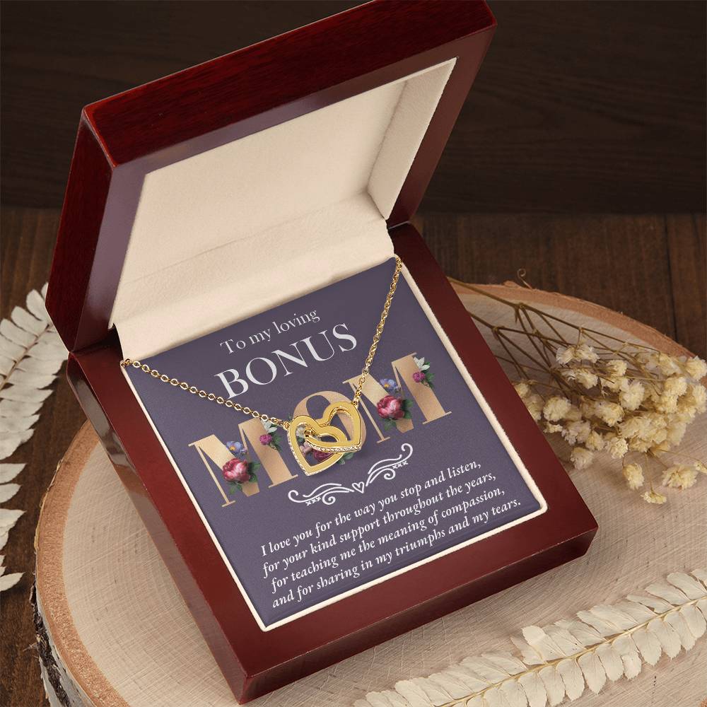 The Bonus Mom-Throughout The Years Interlocking Hearts Necklace, in an 18k yellow gold finish, is presented in an open wooden jewelry box. The lid displays a heartfelt message for a "Bonus Mom," surrounded by dried flowers, making it an ideal token of love and gratitude.