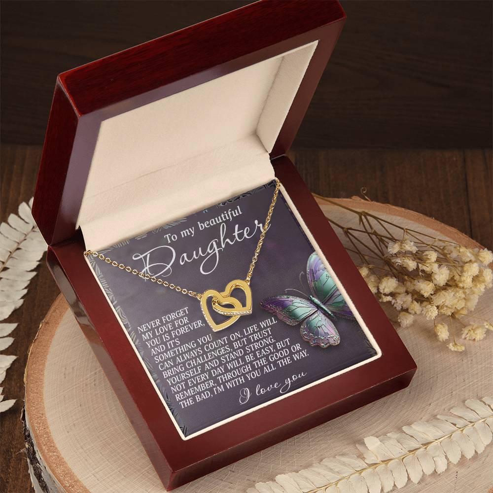 The Daughter-Forever Love Interlocking Hearts necklace in rose gold is showcased on a wooden surface with dried flowers, nestled in an open red box. The lid features a heartfelt message and colorful butterfly image, while cubic zirconia crystals add sparkle to this romantic piece.