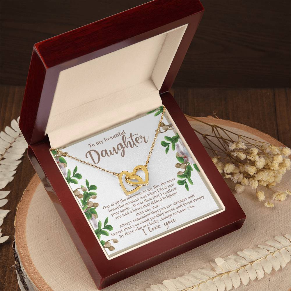 The "Daughter-First Smile" Interlocking Hearts necklace, in rose gold with cubic zirconia, sits in an open red box. Behind it is a floral note to a daughter. The box rests on wood, next to dried flowers.