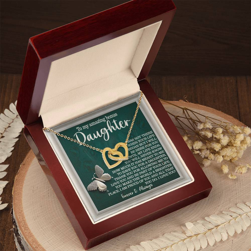 The Daughter-Your Life - Interlocking Hearts Necklace, featuring an angel wing charm and cubic zirconia crystals, is displayed in a wooden jewelry box with a heartfelt message for a daughter. The box sits on a circular wooden surface adorned with foliage.