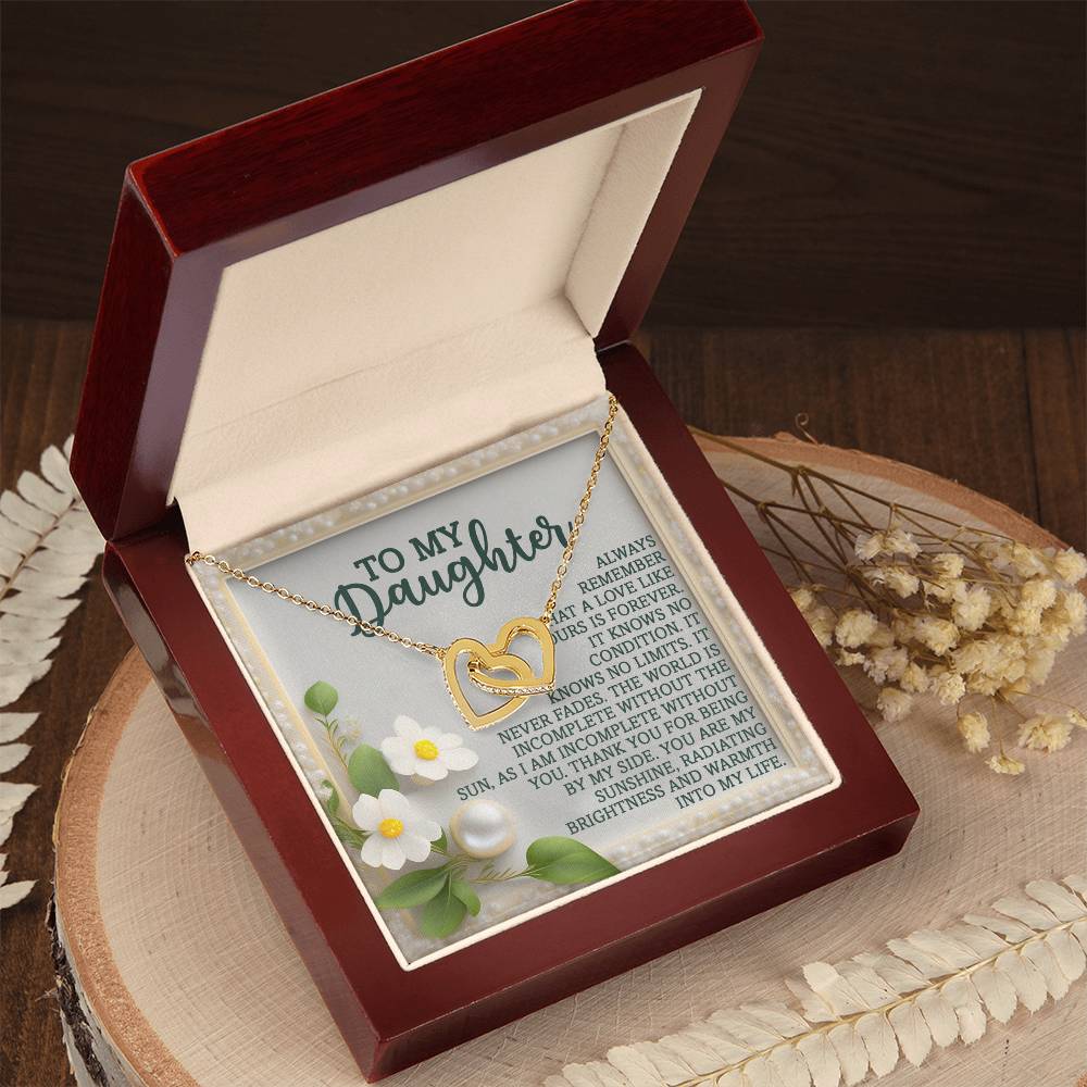 The Daughter-Love Forever - Interlocking Hearts Necklace, with a rose gold finish, gleams in an open velvet-lined red box featuring a heartfelt "To My Daughter" message and floral design. Nestled on a wooden surface, it is adorned with nearby dried flowers.