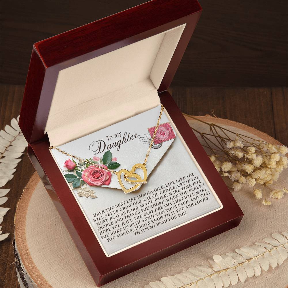 The Daughter-My Wish Interlocking Hearts Necklace, featuring cubic zirconia crystals in a gold heart shape, rests in a dark red box. A card with "To my Daughter," pink roses, and a heartfelt note accompanies it. The rose gold finish adds elegance on a wooden surface with decorative leaves nearby.