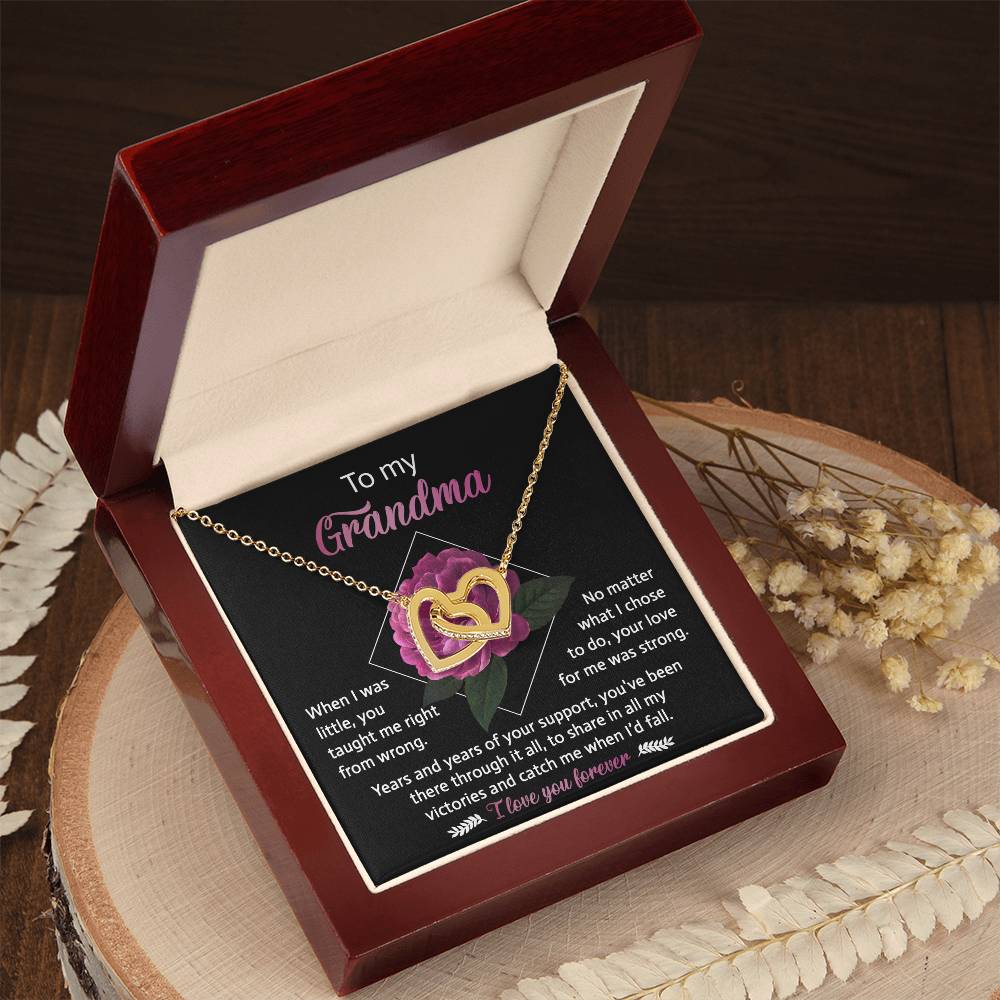 The Grandma-Love For Me Interlocking Hearts Necklace with rose gold finish, adorned with cubic zirconia and decorative leaves and flowers, rests in a wooden box featuring a loving message for grandmother's supportive wisdom.