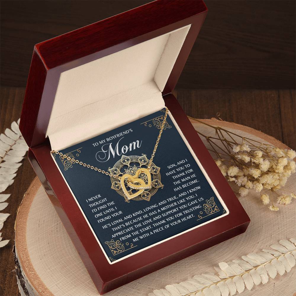 On a wooden surface, a jewelry box holds the Mom-Thankful Heart - Interlocking Hearts necklace in rose gold. The lid reads "To My Boyfriend's Mom" with a heartfelt message, complemented by dried flowers.