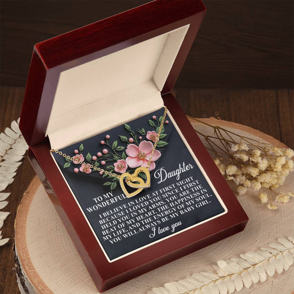 A wooden box holds the Daughter-Love Sight - Interlocking Hearts Necklace, featuring a gold heart pendant with pink flower accents. Inside the lid, a sentimental message is displayed in white text. The scene is completed by dried flowers near the box on the wooden table.
