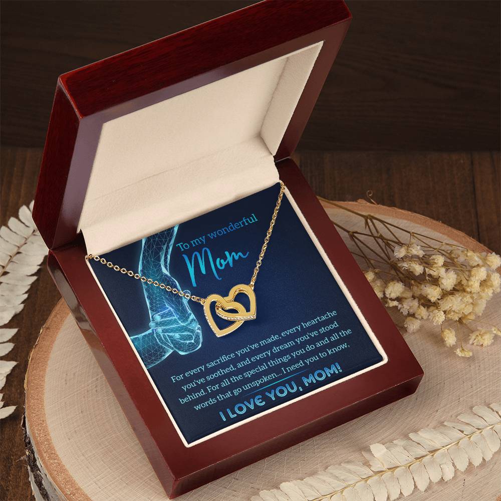 A personalized gift featuring the "To Mom, Stood Behind - Interlocking Hearts Necklace" with cubic zirconia crystals, elegantly presented in a red velvet jewelry box and accompanied by a heartfelt card for Mom, resting on a wooden surface surrounded by delicate dried flowers.