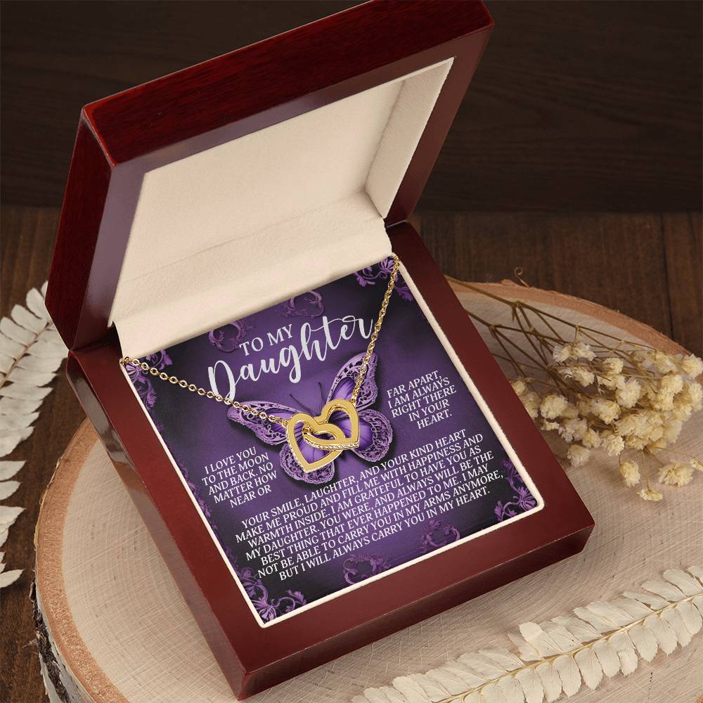 An open jewelry box presents the Interlocking Hearts necklace, Daughter-Moon Back edition, in rose gold on a "To My Daughter" card with a heartfelt message. It sits on a wooden surface decorated with dried flowers and sparkling cubic zirconia crystals.
