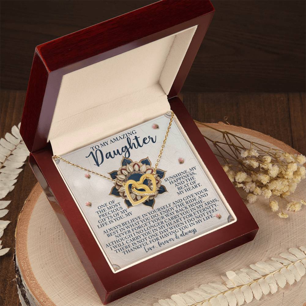 An open jewelry box reveals the Daughter-Precious Gift - Interlocking Hearts Necklace with a rose gold finish. Inside is a heartfelt letter titled "To My Amazing Daughter," expressing love and admiration. The box sits on a wooden surface adorned with decorative dried flowers.