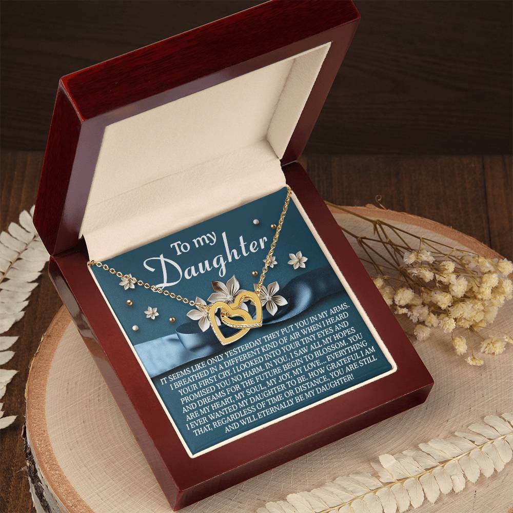 The Daughter-Hopes Dreams - Interlocking Hearts Necklace, a gold heart-shaped pendant with cubic zirconia crystals, sits in an open red box reading "To my Daughter." It's set on wood with decorative flowers, captivating with its elegant design.
