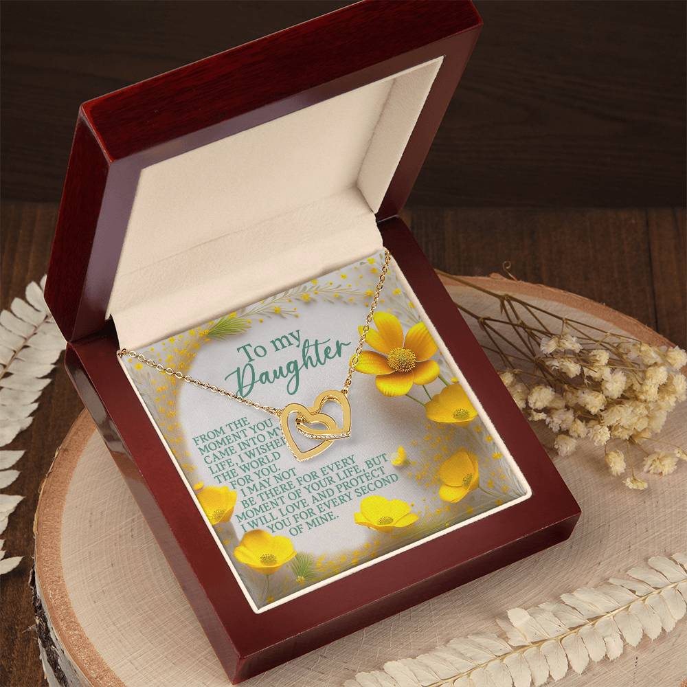 The Daughter-Every Moment - Interlocking Hearts Necklace, featuring a heart-shaped pendant encrusted with cubic zirconia, is elegantly presented in an open wooden box. Inside, yellow flowers frame a loving message to a daughter. The box is displayed on wood and adorned with dried flowers.
