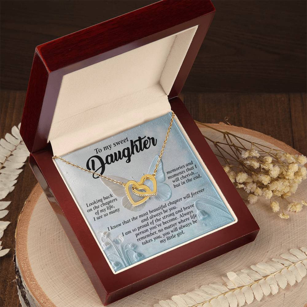 An open jewelry box reveals the "Daughter-Beautiful Chapter" Interlocking Hearts necklace, crafted in gold and embellished with cubic zirconia. Inside the lid, a heartfelt message of love and pride for a daughter is displayed. Dried flowers encircle the box on a wooden surface.