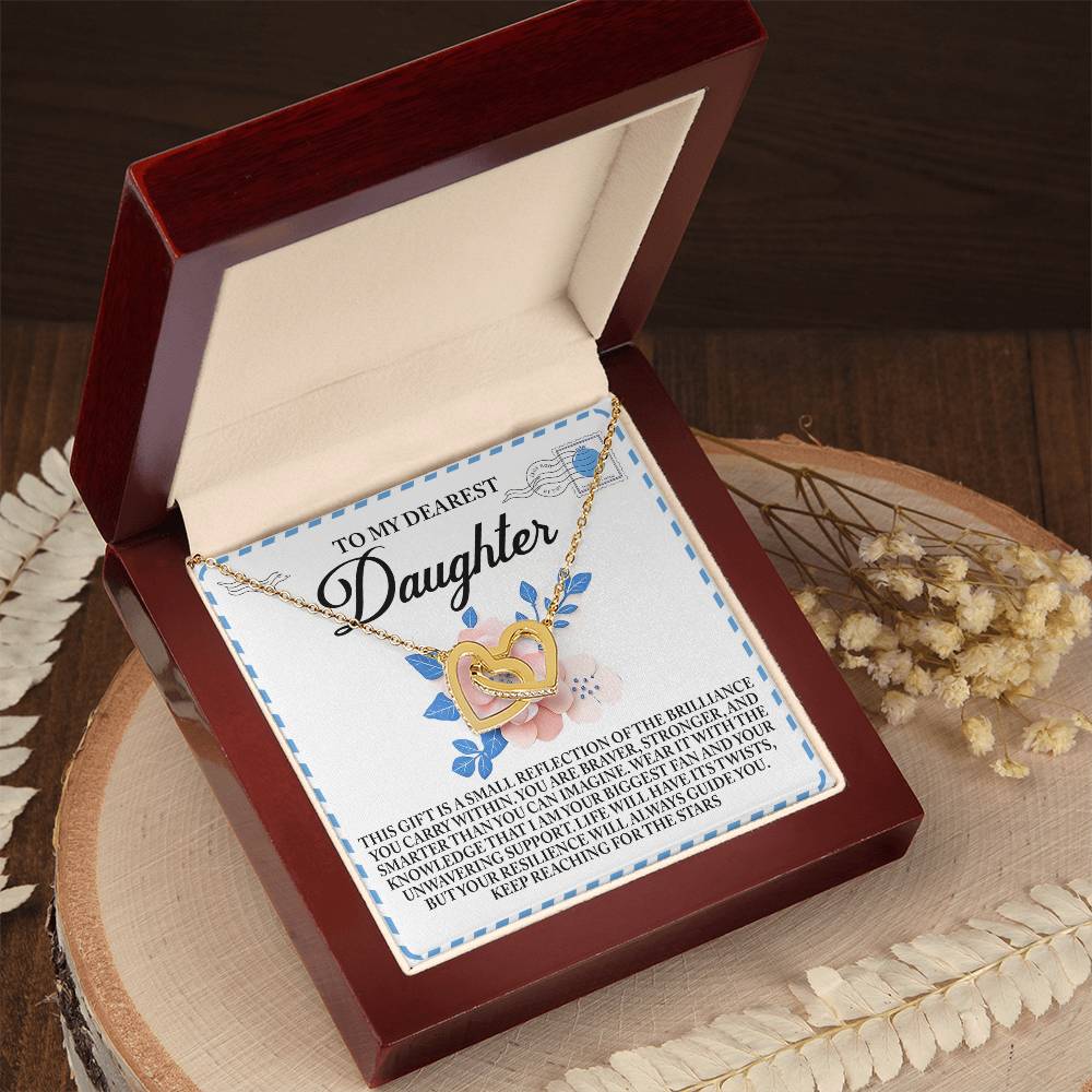 The Daughter-Brilliance Within - Interlocking Hearts Necklace, crafted from two-tone surgical steel and gold, is presented in an open jewelry box inscribed with "To My Dearest Daughter." Dried flowers nearby enhance its sentimental charm on the wooden surface.
