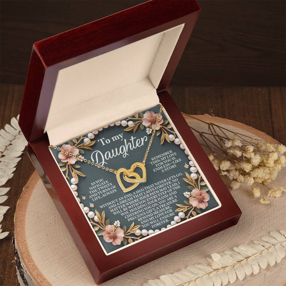 A jewelry box containing the "Daughter-Endless Love - Interlocking Hearts Necklace" features a heart-shaped pendant with cubic zirconia, set against a rose gold-accented interior lid with floral and pearl-like designs, resting on wood beside dried flowers.