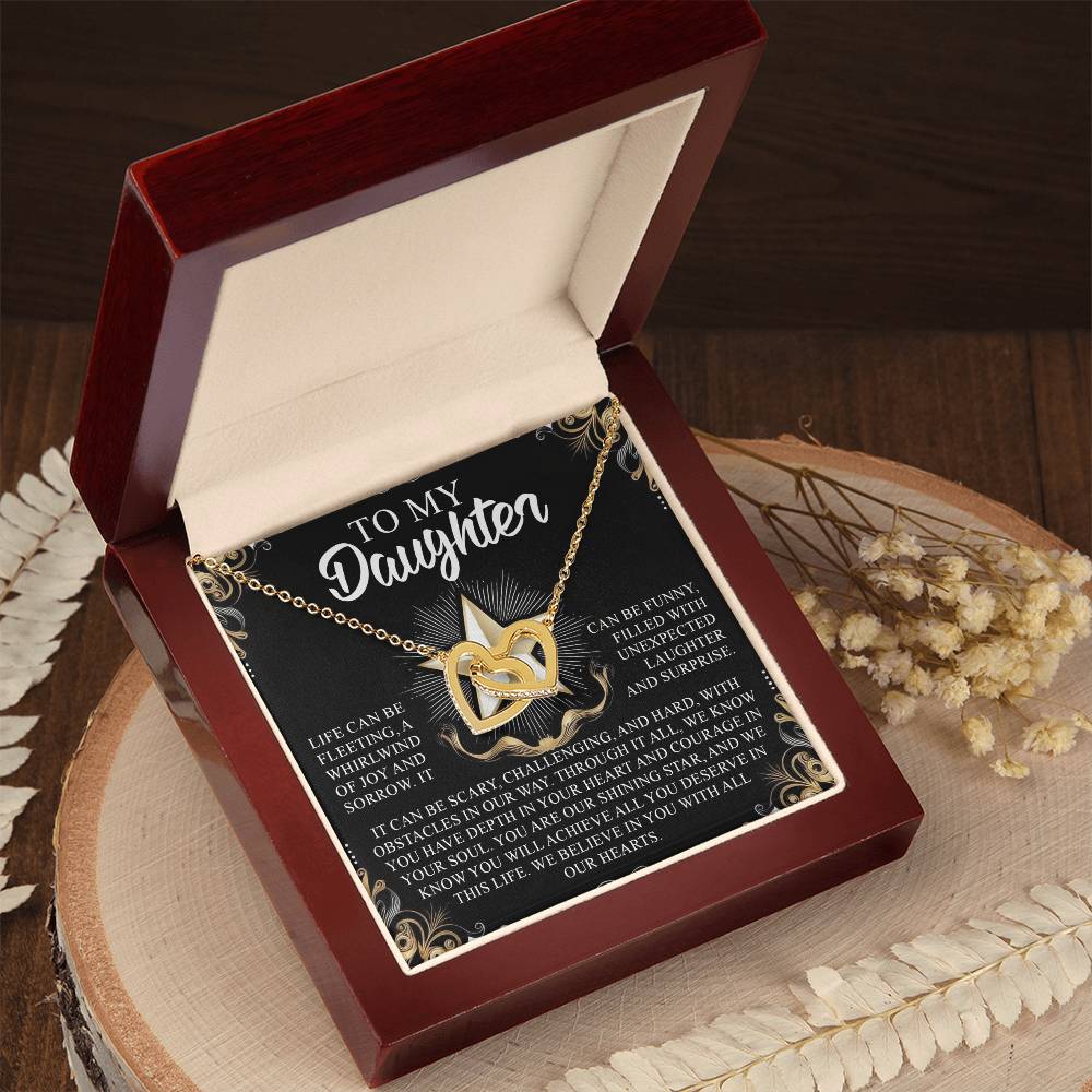 The Daughter-Shining Star Interlocking Hearts Necklace, with a rose gold finish, is presented in an open wooden box. It includes a sentimental message card stating "To my daughter." The note, elegantly bordered, lies on a round wooden surface decorated with dried flowers.