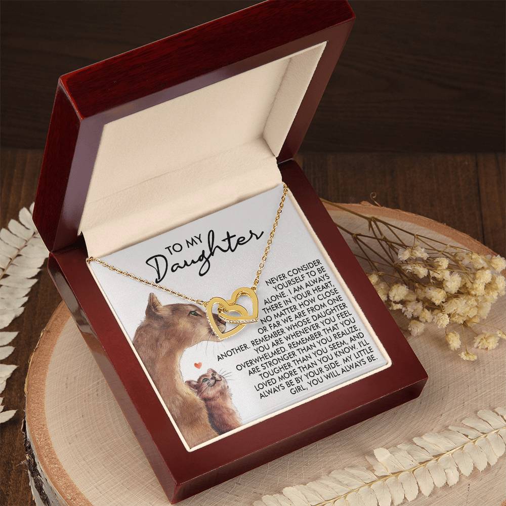 An Interlocking Hearts Necklace in 18k yellow gold finish, named "Daughter-Never Alone," is displayed in an open wooden jewelry box. Inside the lid, a touching "To My Daughter" message accompanies a lion and cub illustration. The box sits on a round wooden surface, accented by dried flowers.