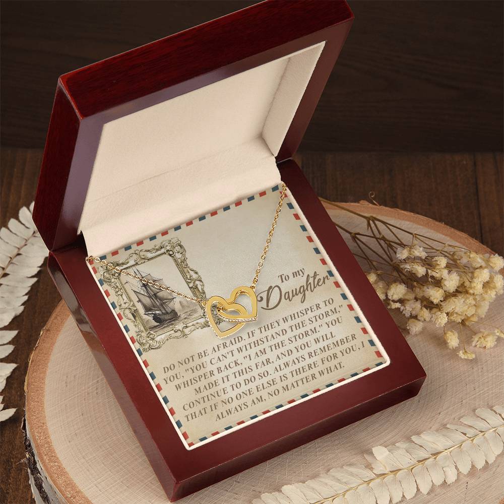 A luxury box unveils the Daughter-Withstand Storm Interlocking Hearts Necklace, featuring a gold heart-shaped design with cubic zirconia crystals. It rests on a heartfelt card to a daughter, displayed on wood and surrounded by white dried flowers.