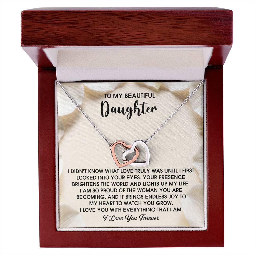 The Daughter-Into Your Eyes - Interlocking Hearts Necklace, adorned with shimmering cubic zirconia crystals, rests elegantly in a jewelry box. It features an inscription about love and pride for a daughter, beautifully accentuated by its delicate rose gold finish.