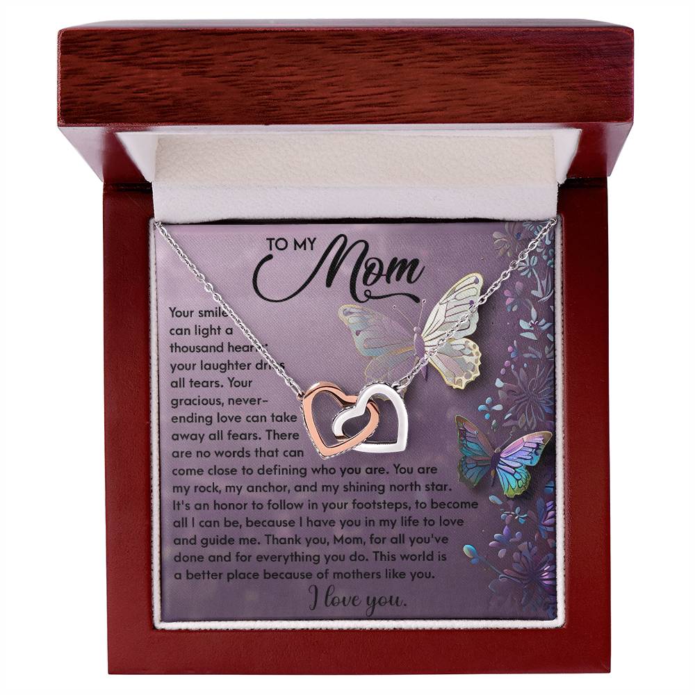 The Mom-Shining Star - Interlocking Hearts Necklace, adorned with cubic zirconia crystals, nestles in a luxury box featuring a heartfelt message to "Mom" surrounded by vibrant butterflies and floral patterns.