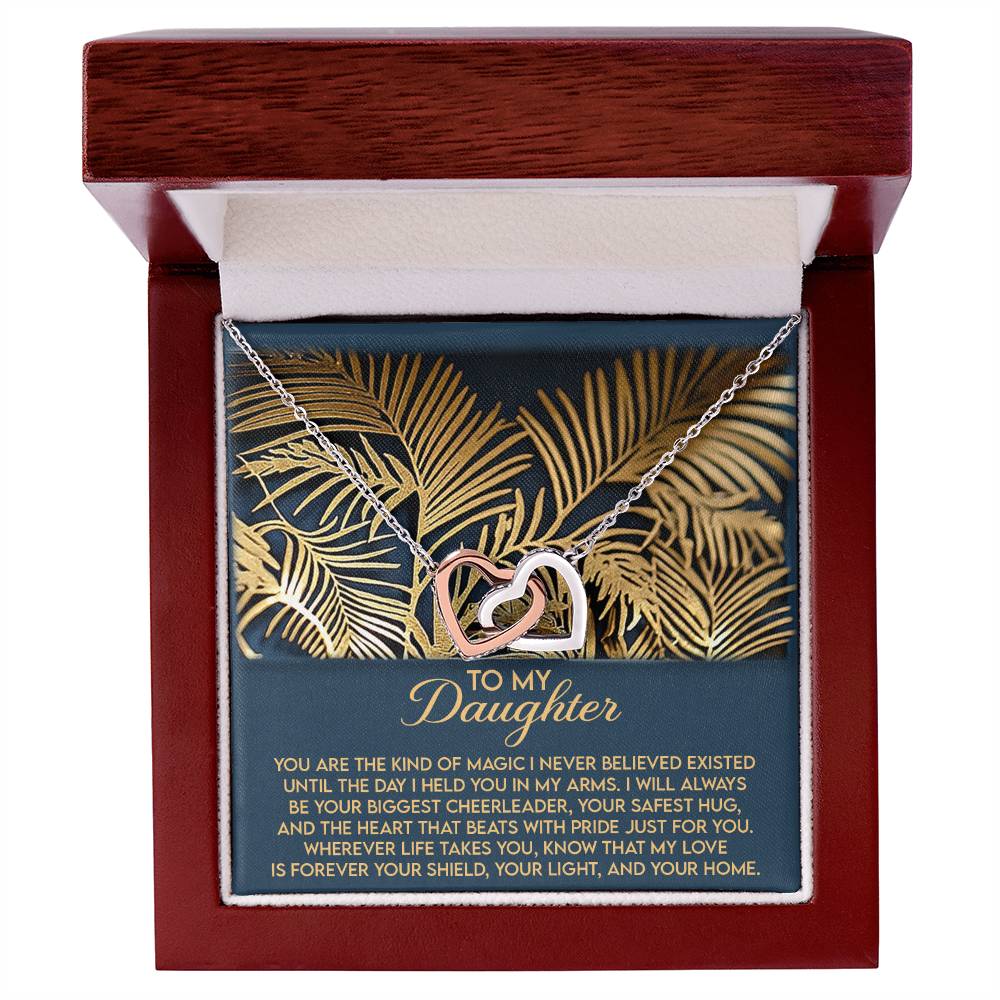 The Daughter-Your Home Interlocking Hearts Necklace, featuring a rose gold finish and cubic zirconia crystals, is beautifully presented in a wooden box with a gold leaf design on a lush green background—an elegant and heartfelt gift for your daughter.