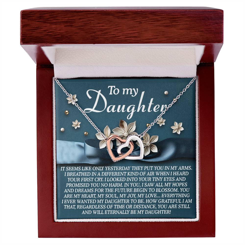The Daughter-Hopes Dreams Interlocking Hearts Necklace, with a silver and rose gold finish adorned with delicate flowers, is elegantly displayed in a jewelry box. Inside, a heartfelt message dedicated "To my Daughter" expresses unwavering love and pride.
