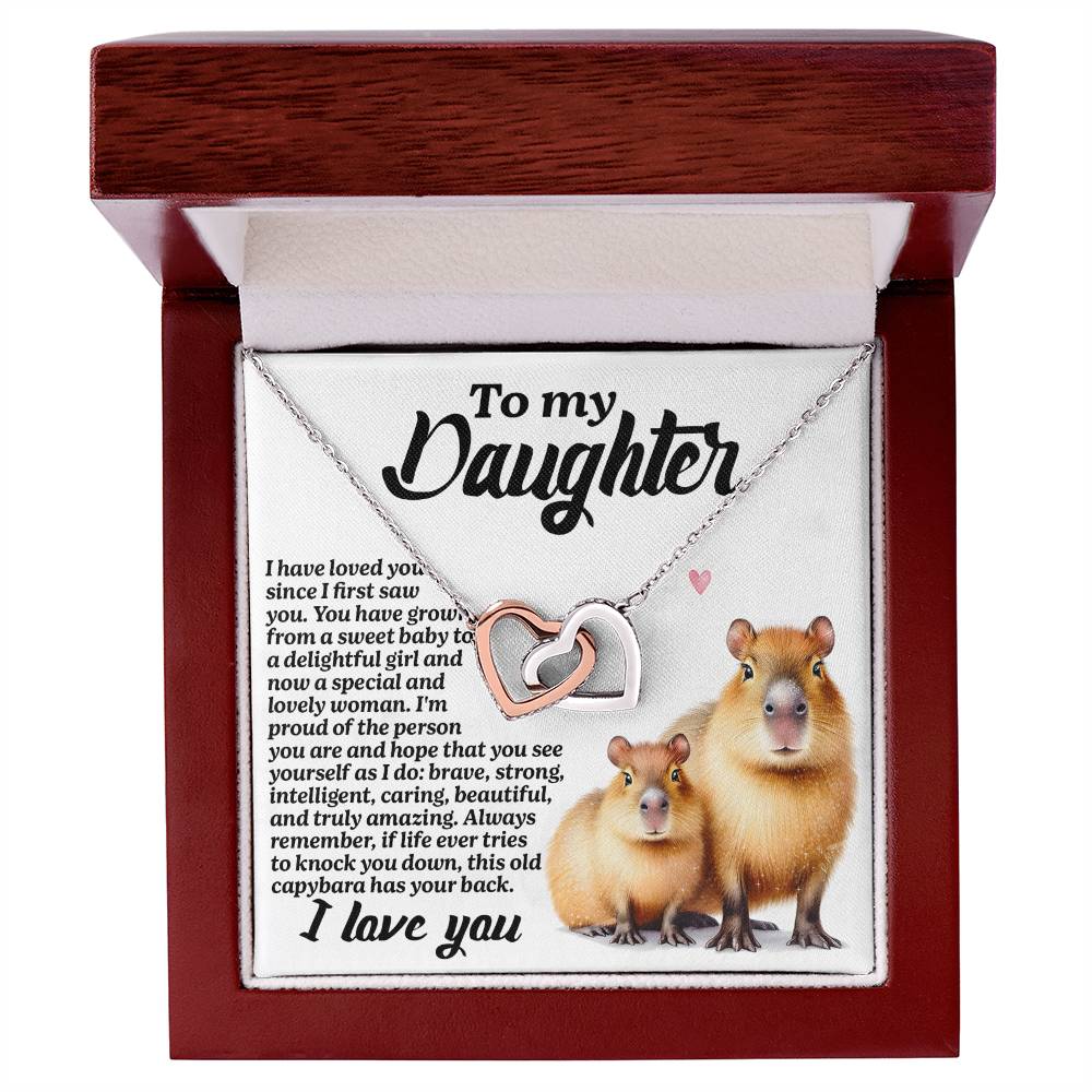 The Daughter-Sweet Baby - Interlocking Hearts Necklace features intertwined hearts adorned with cubic zirconia crystals and is finished in surgical steel and rose gold. The box reads "To my Daughter," includes a heartfelt message, and features an illustration of two capybaras.
