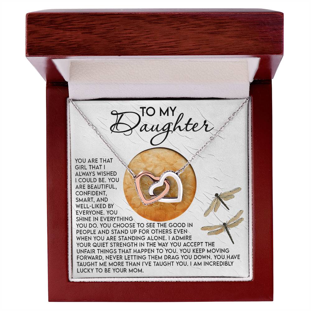 The Daughter-Quiet Strength Interlocking Hearts Necklace, with a rose gold finish and cubic zirconia crystals, is elegantly displayed in an open wooden box. A heartfelt message beginning with "To my daughter" graces the card, beautifully adorned with dragonfly illustrations.