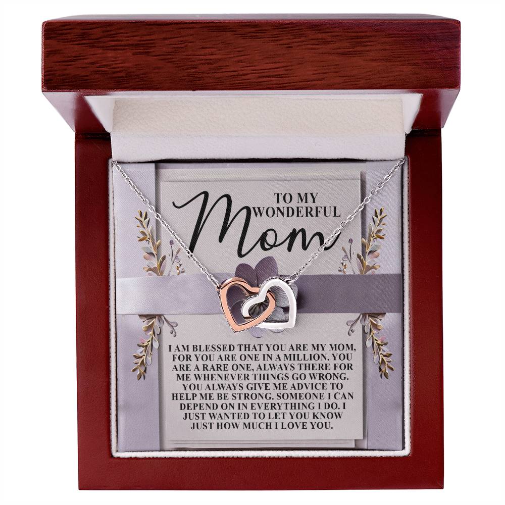 The Mom-Rare One - Interlocking Hearts Necklace, with one heart in silver and the other in rose gold adorned with cubic zirconia crystals, is presented in an open wooden box. A card behind it reads "To My Wonderful Mom" surrounded by floral accents.
