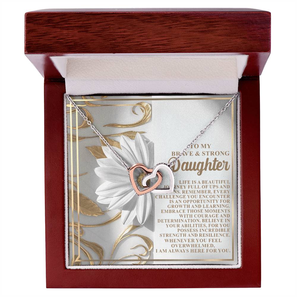 The Daughter-Here For You Interlocking Hearts Necklace in silver and rose gold finish is elegantly presented in a wooden box. Behind it, a heartfelt message for a daughter, with floral artwork and cubic zirconia crystals, expresses love and encouragement.