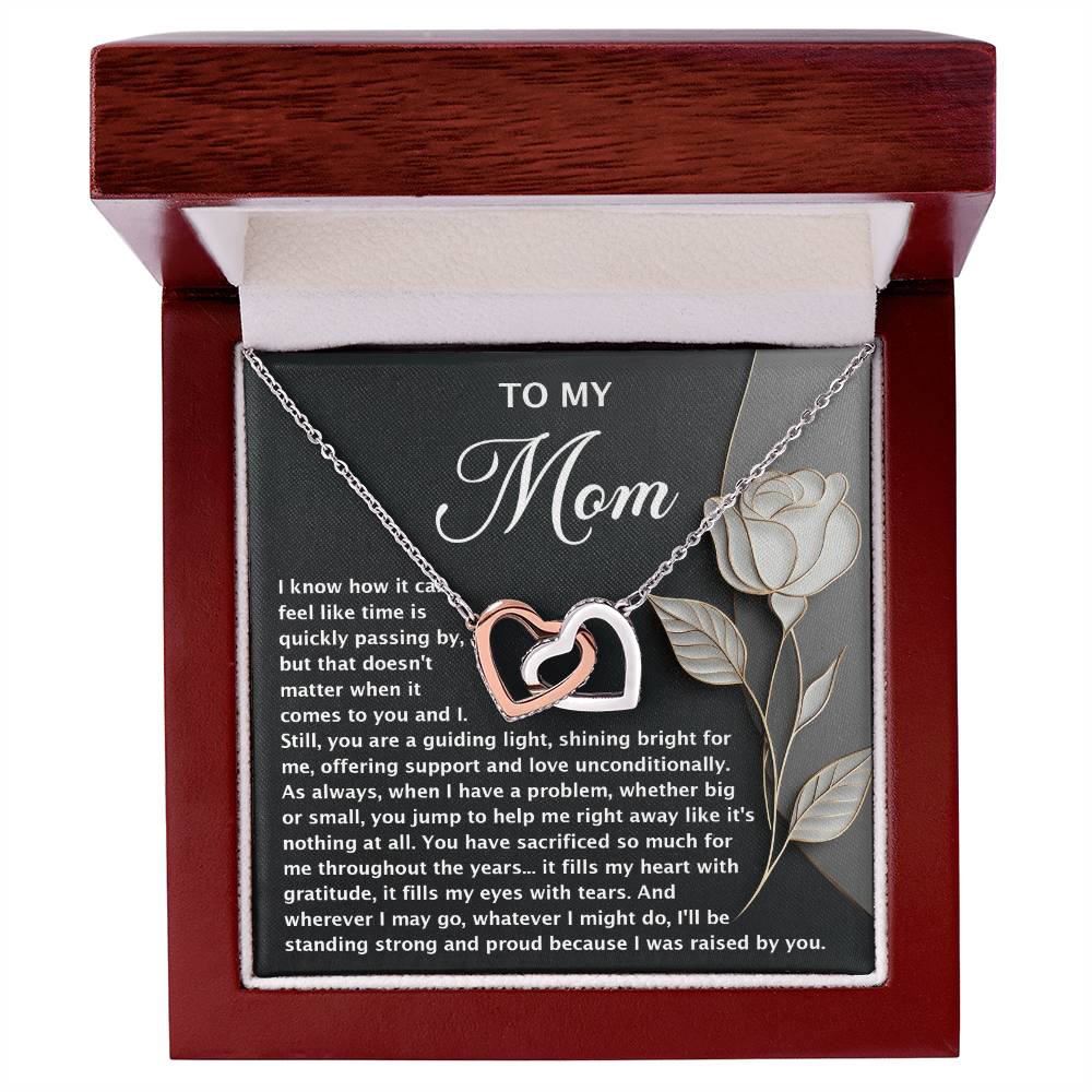 The Mom-Guiding Light Interlocking Hearts Necklace, with rose gold finish, is elegantly displayed in a wooden box. Its lid features a heartfelt message for "Mom" and a white rose illustration, while the soft interior lining gently cradles the exquisite piece.
