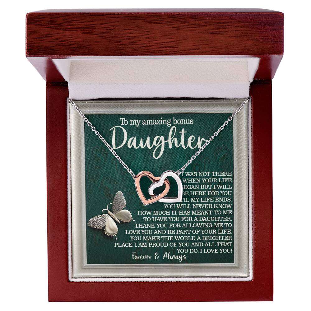 A polished wooden jewelry box holds the "Daughter-Your Life" Interlocking Hearts necklace, featuring shimmering cubic zirconia crystals in a rose gold finish and a loving message for a "bonus daughter," expressing love and pride.