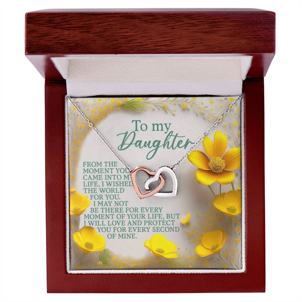 The Daughter-Every Moment - Interlocking Hearts Necklace, made from surgical steel and cubic zirconia crystals, is presented in a wooden box. Yellow flowers in the background enhance the "To my Daughter" message, symbolizing love and protection.
