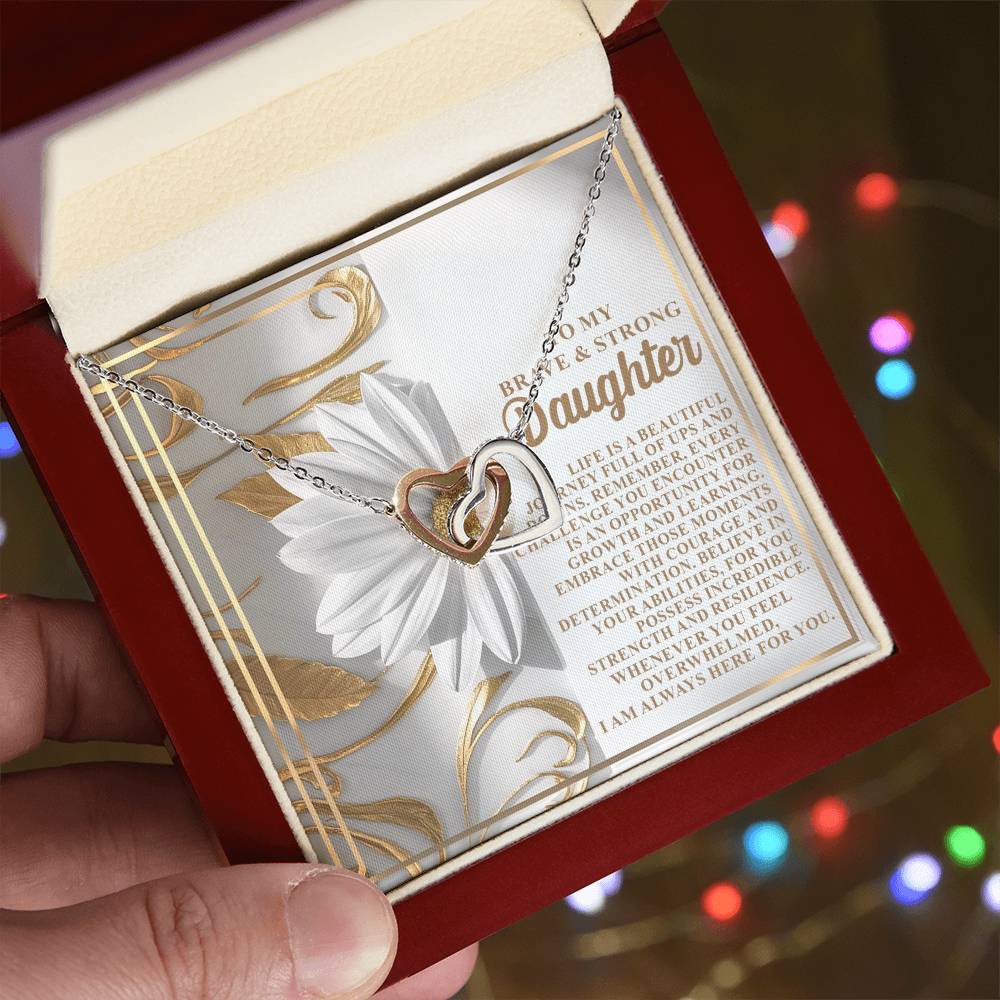 A hand holds a red box showcasing the Daughter-Here For You - Interlocking Hearts Necklace, adorned with cubic zirconia crystals. Behind it is a heartfelt card for a daughter, set against a softly glimmering backdrop of colorful lights.