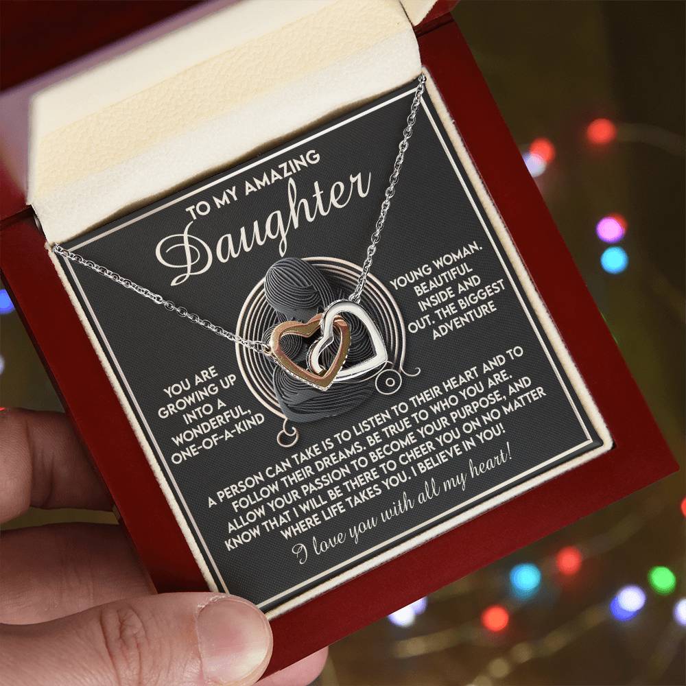 A person holds an open maroon gift box with a white interior, containing the "Daughter-Your Dreams - Interlocking Hearts Necklace," featuring a heart-shaped pendant adorned with cubic zirconia crystals. In the background is a heartfelt message expressing love and admiration for a daughter.