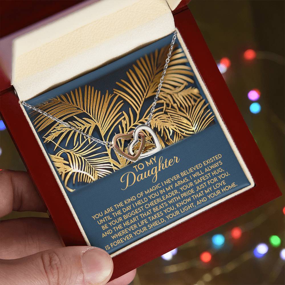 A hand holds an open jewelry box showing the "Daughter-Your Home - Interlocking Hearts Necklace" with a silver heart and cubic zirconia crystals. A card with gold palm leaf designs carries a touching message for a daughter, glowing against colorful blurred lights in the background.