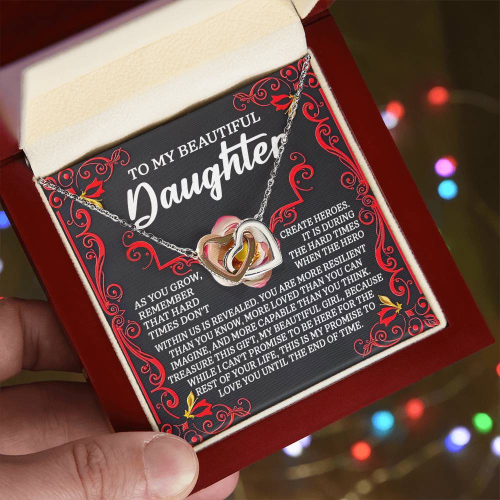 A hand displays an open box with a Daughter-Hard Times - Interlocking Hearts Necklace in rose gold, adorned with cubic zirconia crystals and set on a card that says, "To My Beautiful Daughter," surrounded by elegant red patterns.
