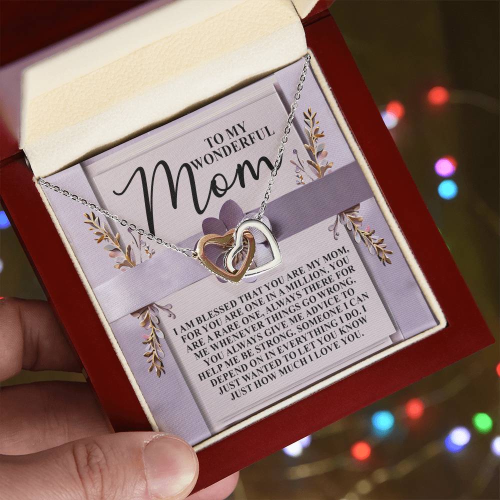 A person holds an open red jewelry box with the stunning Mom-Rare One - Interlocking Hearts Necklace made from surgical steel and adorned with cubic zirconia crystals, nestled against a heartfelt "Mom" card. In the background, colorful lights gently twinkle.
