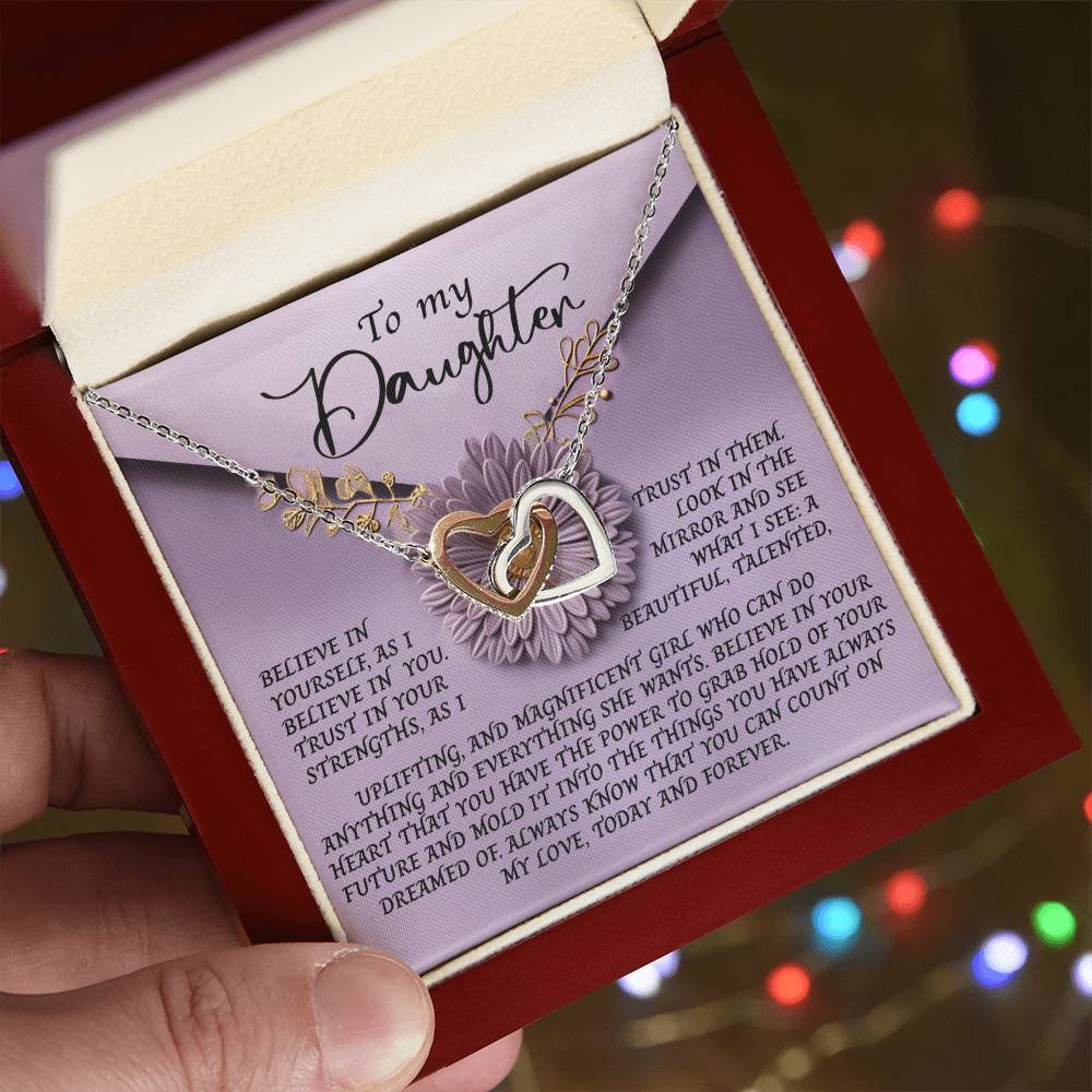 A hand reveals an open red box containing the Daughter-Magnificent Girl - Interlocking Hearts Necklace, showcasing intertwined gold and silver hearts adorned with cubic zirconia. The background features a heartfelt card for a daughter set against blurred colorful lights.