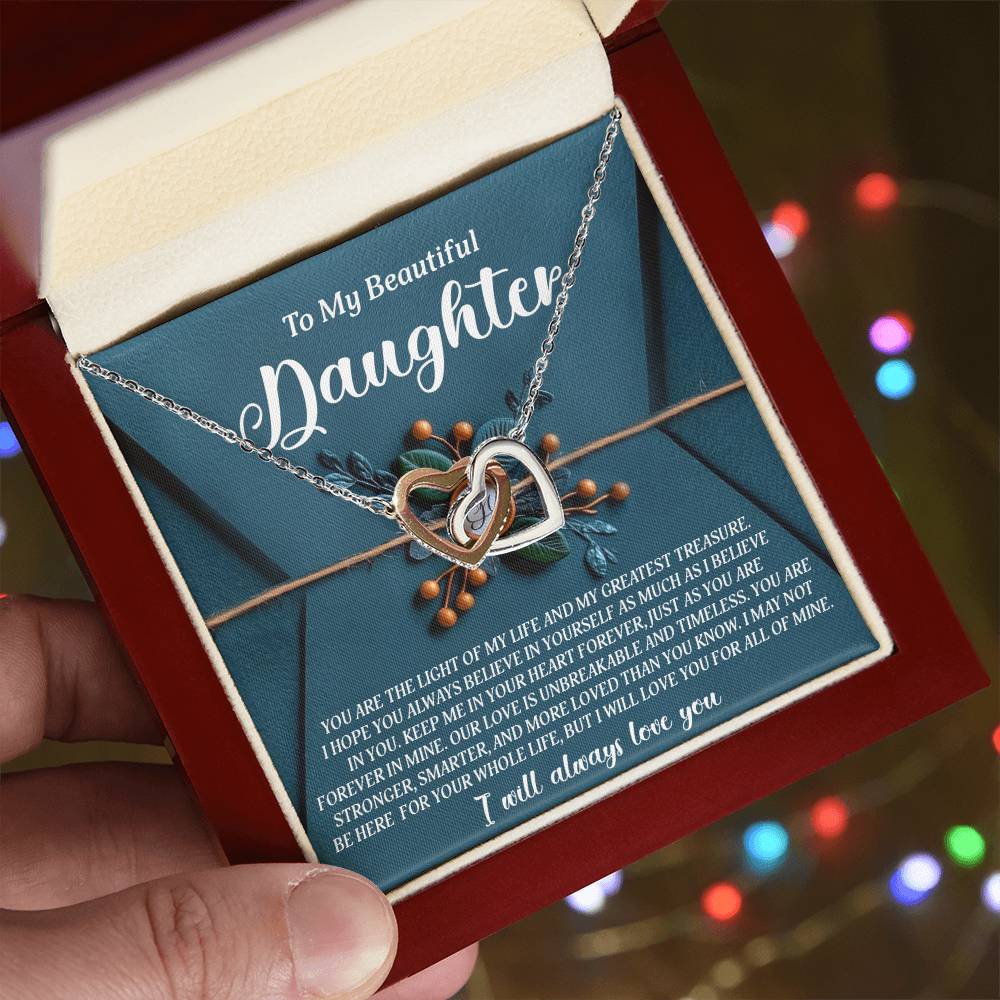 A hand holds an open red jewelry box displaying the "Daughter-All Of Mine - Interlocking Hearts Necklace" with cubic zirconia crystals. Inside, a message starts with "To My Beautiful Daughter," featuring a heartfelt note of admiration and love, as soft, multicolored lights blur in the background.