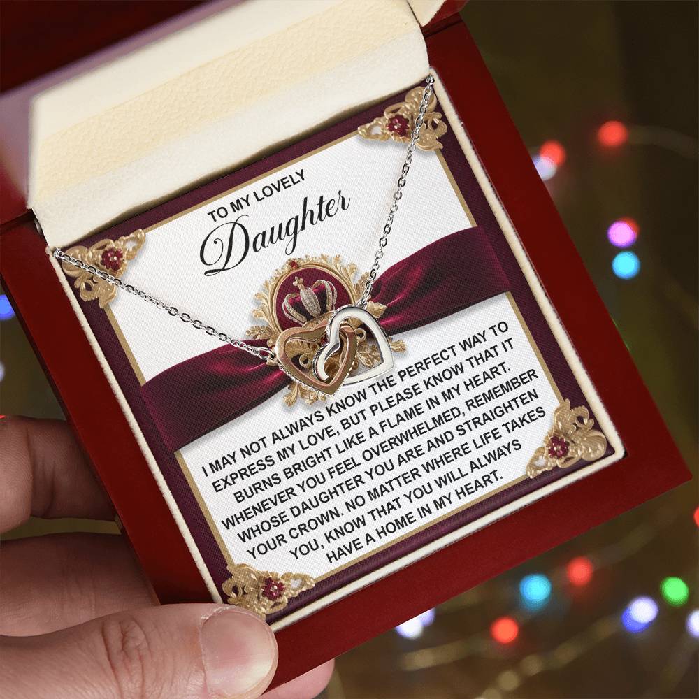 A hand holds a gift box with the Daughter-A Flame Interlocking Hearts necklace, featuring cubic zirconia crystals. The rose gold finish sparkles beautifully against blurred, colorful lights alongside a heartfelt message for a daughter.