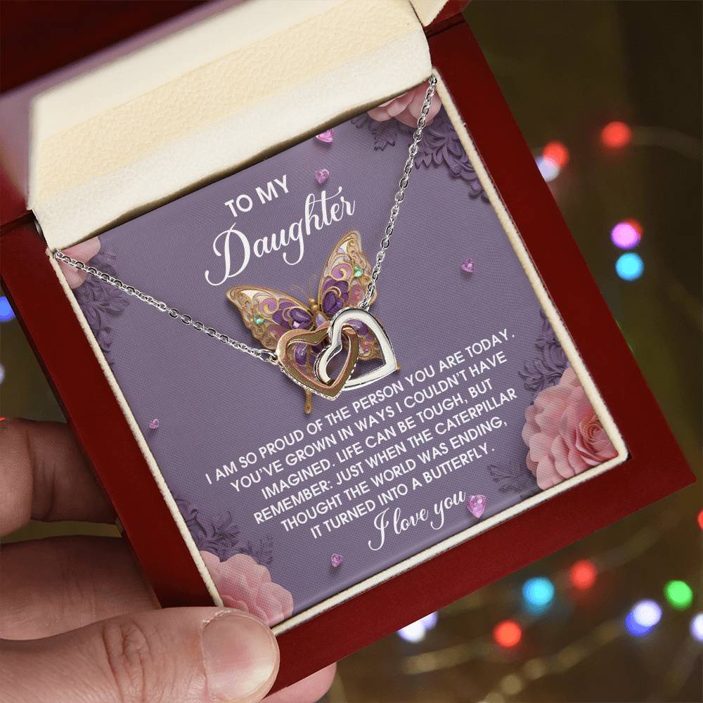 A person holds an open jewelry box with the Daughter-A Butterfly Interlocking Hearts Necklace, adorned with cubic zirconia crystals and a rose gold finish. An inscription for their daughter shimmers in the light against a softly glowing background of blurred lights.