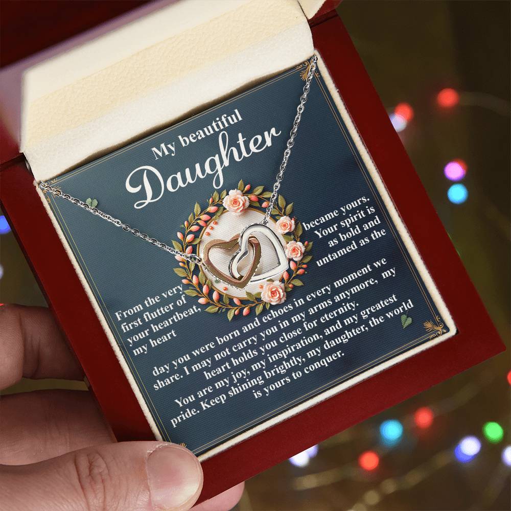 A hand holds an open red box displaying the Daughter-First Flutter - Interlocking Hearts Necklace in rose gold, adorned with sparkling cubic zirconia. A heartfelt "My Beautiful Daughter" message sits amidst decorative flowers, with blurred colorful lights in the background.