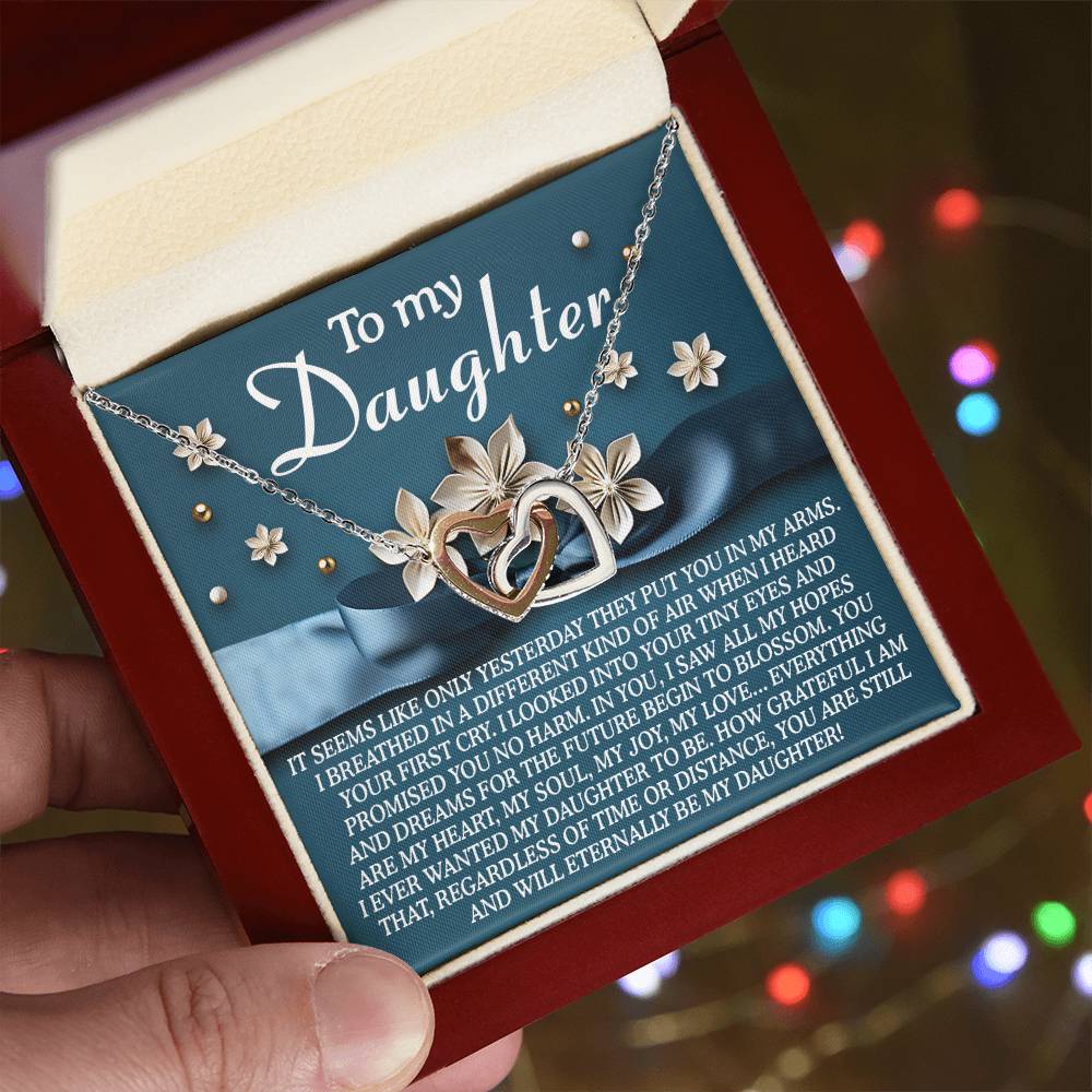 A hand presents a red jewelry box with the Daughter-Hopes Dreams - Interlocking Hearts Necklace. The silver necklace features floral details and a rose gold finish, casting hues over the cubic zirconia crystals. An emotional message for a daughter resonates softly in the blurred colorful lights.