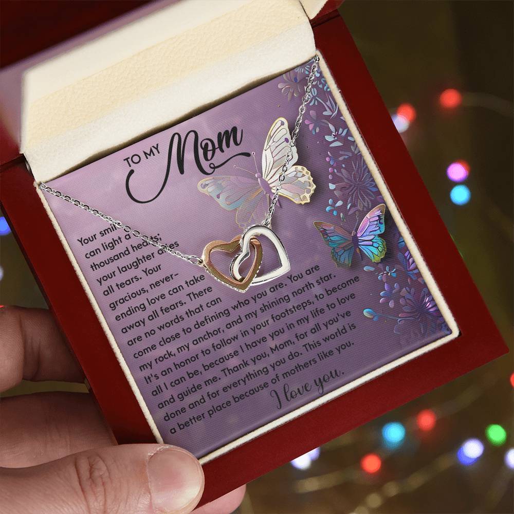 A hand opens a luxury box displaying the Mom-Shining Star - Interlocking Hearts necklace with cubic zirconia crystals. The lid shows a heartfelt message to "Mom" amid colorful butterflies and floral designs on a purple background, with softly blurred festive lights in the distance.
