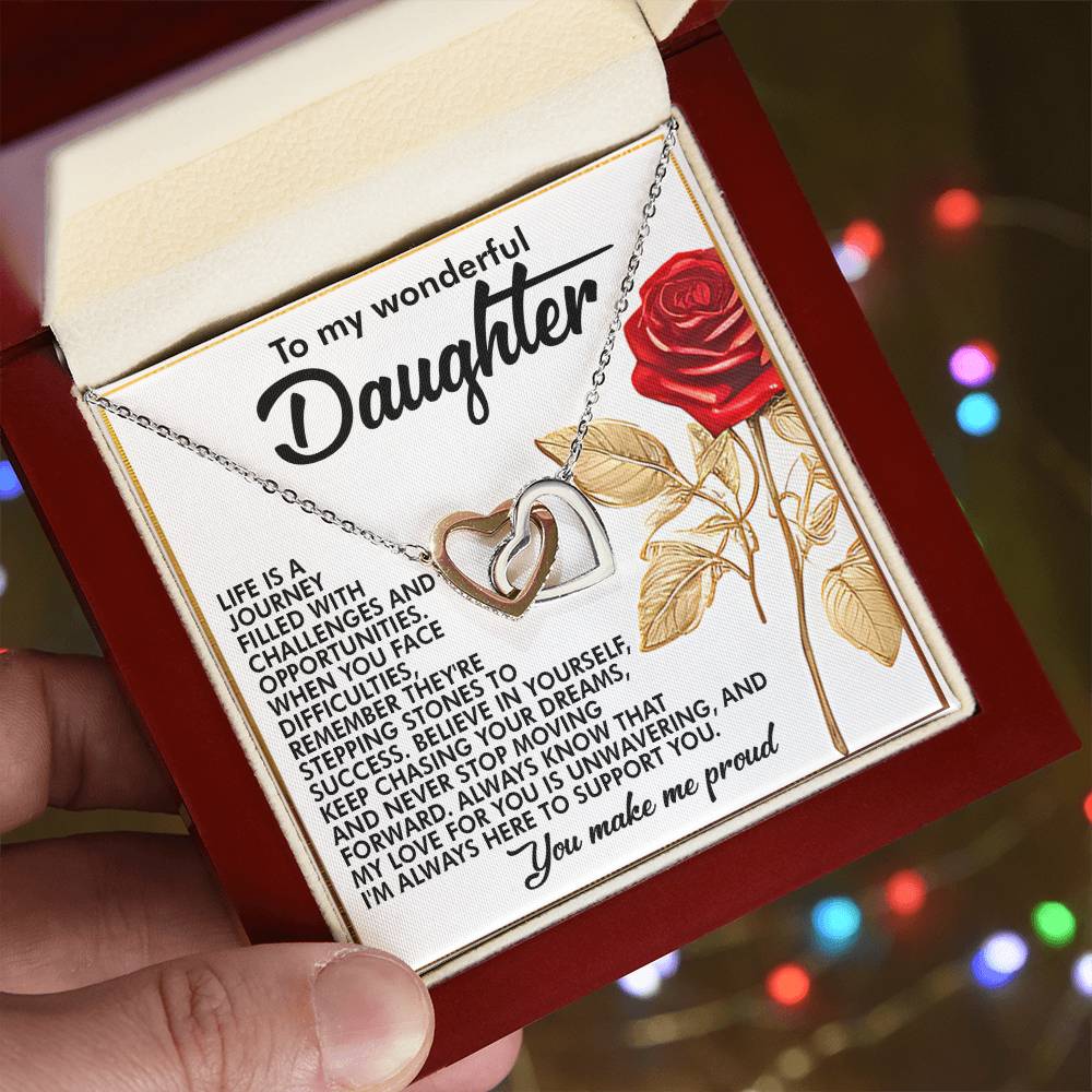 A person holds an open jewelry box with the Daughter-Journey Challenges - Interlocking Hearts Necklace in rose gold. The lid has appreciative words for a daughter and a red rose illustration, against a softly blurred background with multicolored lights.