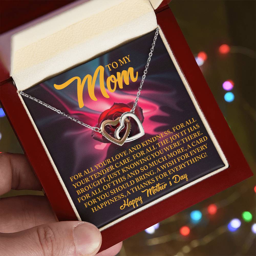 A person holds an open red jewelry box showcasing the Mom-Tender Care - Interlocking Hearts Necklace with cubic zirconia crystals. The lid bears a heartfelt Mother's Day message of love and gratitude, with softly twinkling blurred colorful lights in the background enhancing the sentiment.