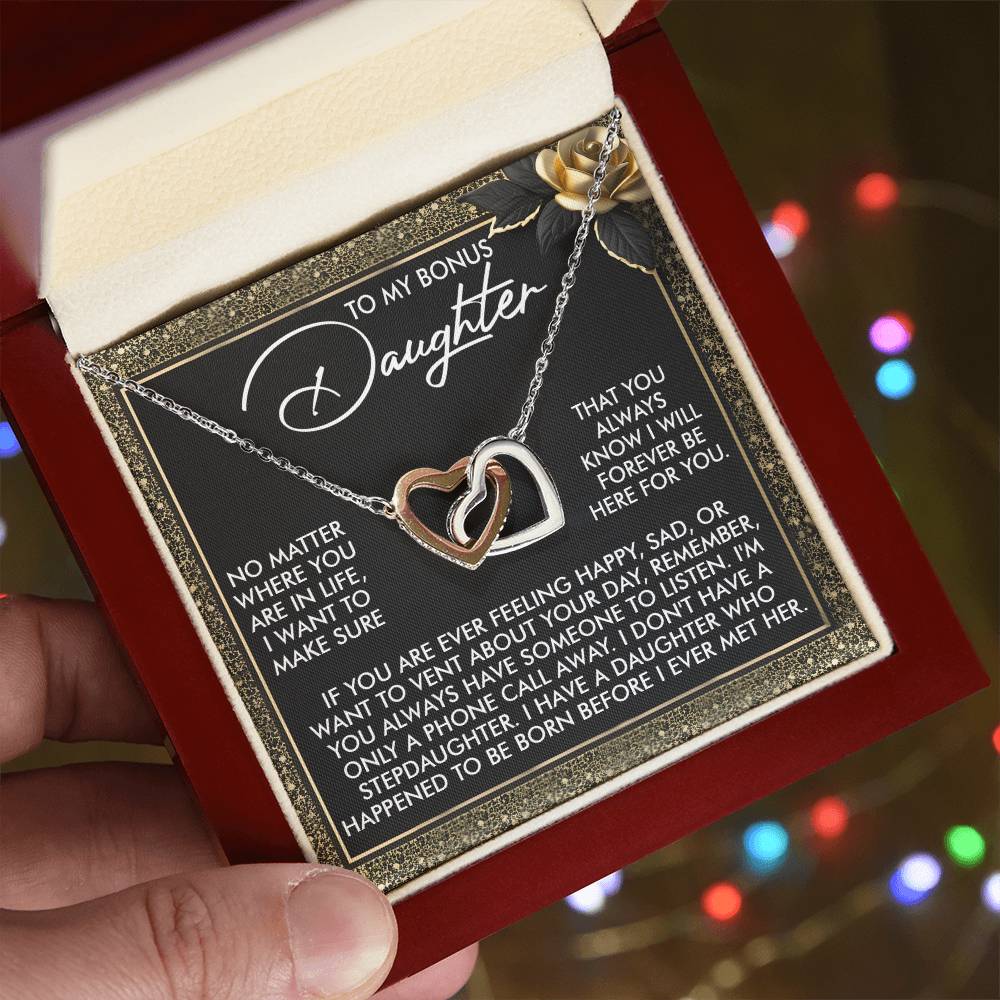 A hand holds an open jewelry box with the Daughter-Phone Call - Interlocking Hearts Necklace made of surgical steel and cubic zirconia. The lid's interior displays a heartfelt message titled "To My Bonus Daughter" on a black and gold background, accented by colorful blurred lights.