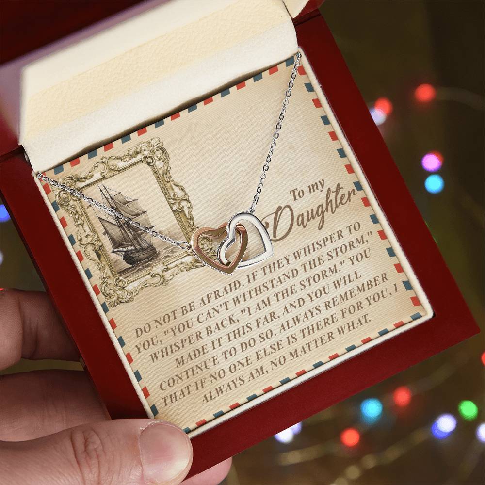 A hand holds the Daughter-Withstand Storm - Interlocking Hearts Necklace, finished in stunning rose gold. The background features a card with an old sailing ship and a motivational message about resilience to a daughter, enhanced by sparkling cubic zirconia crystals.